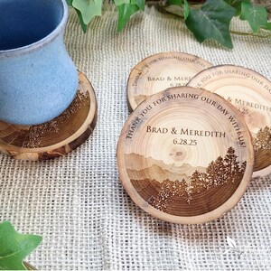 May include: Round wooden coasters featuring a laser-engraved mountain and forest landscape. The text reads, "THANK YOU FOR SHARING OUR DAY WITH US! BRAD & MEREDITH 6.28.25". One coaster supports a light blue ceramic mug, while two others sit nearby.