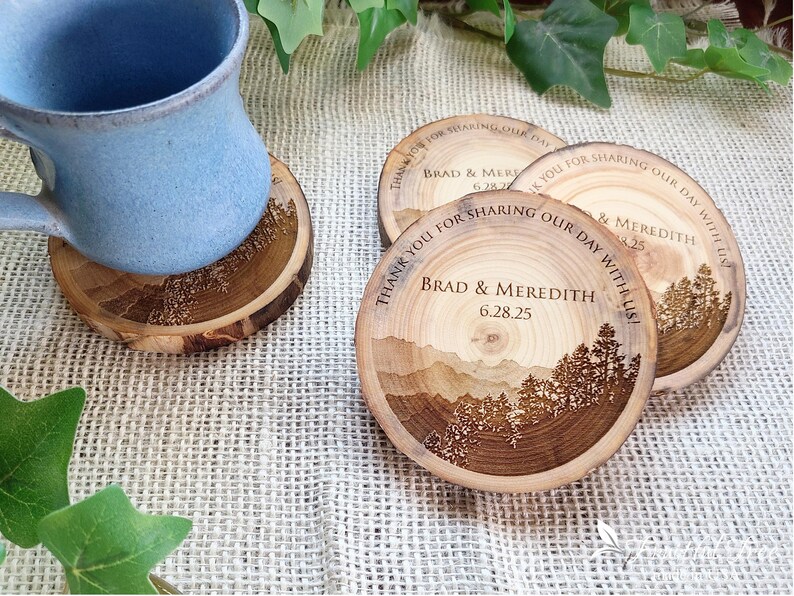 May include: Round wooden coasters featuring a laser-engraved mountain and forest landscape. The text reads, "THANK YOU FOR SHARING OUR DAY WITH US! BRAD & MEREDITH 6.28.25". One coaster supports a light blue ceramic mug, while two others sit nearby.