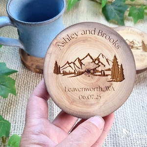 May include: A round wooden coaster featuring a laser-engraved mountain landscape with pine trees. The text reads Ashley and Brooks, Leavenworth, WA, and 06.07.25. The natural wood grain is visible, and the coaster has a dark, burnt-in edge finish.