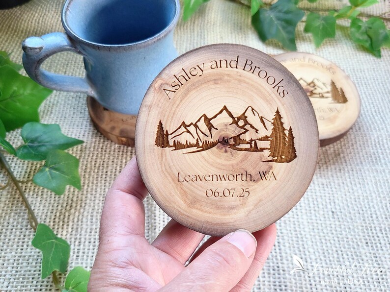 May include: A round wooden coaster featuring a laser-engraved mountain landscape with pine trees. The text reads Ashley and Brooks, Leavenworth, WA, and 06.07.25. The natural wood grain is visible, and the coaster has a dark, burnt-in edge finish.