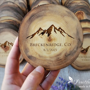 May include: A round wooden slice coaster featuring a dark brown laser-engraved mountain range graphic. The text "BRECKENRIDGE, CO" and "4/1/2023" is centered below the mountains. The wood grain is visible with a natural bark edge.