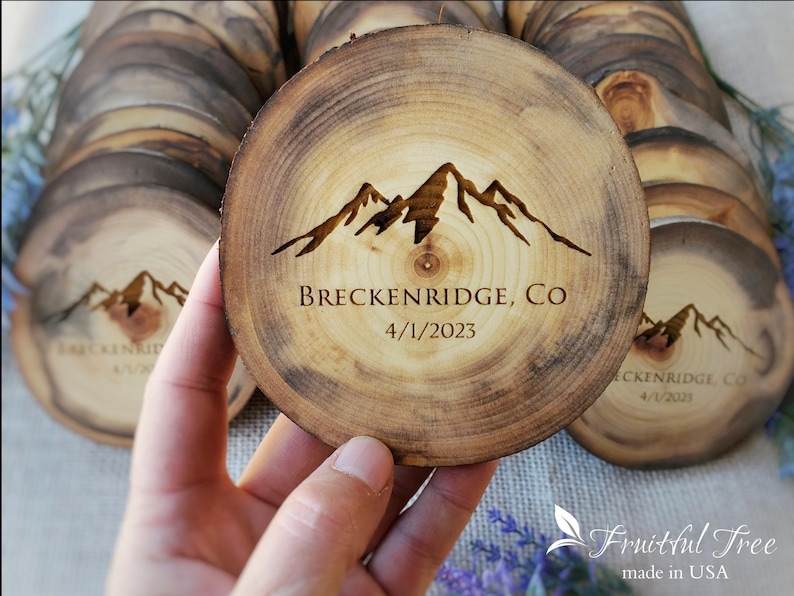 May include: A round wooden slice coaster featuring a dark brown laser-engraved mountain range graphic. The text "BRECKENRIDGE, CO" and "4/1/2023" is centered below the mountains. The wood grain is visible with a natural bark edge.