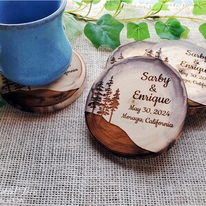 May include: Round wooden coasters with natural bark edges, featuring a laser-engraved pine tree silhouette and the text Sarby & Enrique, May 30, 2024, Moraga, California. A blue ceramic mug rests on one coaster, showcasing the wood grain texture.