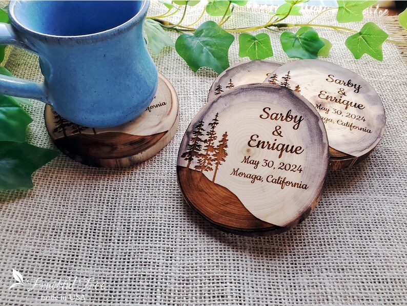May include: Round wooden coasters with natural bark edges, featuring a laser-engraved pine tree silhouette and the text Sarby & Enrique, May 30, 2024, Moraga, California. A blue ceramic mug rests on one coaster, showcasing the wood grain texture.