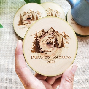 May include: A round wooden coaster featuring a dark brown laser-engraved mountain landscape with pine trees. The text "DURANGO, COLORADO" and "2023" is engraved below the illustration. The coaster has a natural light wood grain and bark edge.