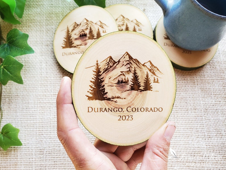 May include: A round wooden coaster featuring a dark brown laser-engraved mountain landscape with pine trees. The text "DURANGO, COLORADO" and "2023" is engraved below the illustration. The coaster has a natural light wood grain and bark edge.