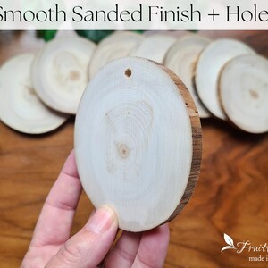May include: A close-up of a smooth, sanded, white wood round with a hole in the centre. The round is being held in a hand. The wood is smooth and has a natural grain pattern. The round is surrounded by other similar rounds.