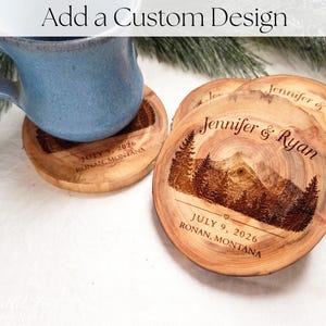 May include: Round wooden coasters featuring a laser-engraved mountain and pine tree landscape. The text reads Jennifer & Ryan, July 9, 2026, and Ronan, Montana. A blue ceramic mug sits on one coaster, and the image includes the text Add a Custom Design.