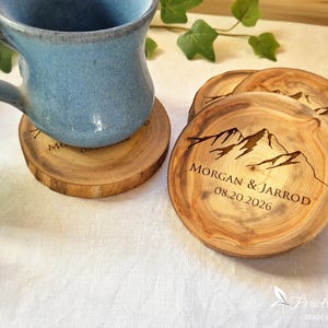 May include: Round wooden coasters with natural bark edges, featuring a laser-engraved mountain range graphic and the text MORGAN & JARROD 08.20.2026. A blue ceramic mug sits on one coaster, while two additional coasters are stacked nearby.