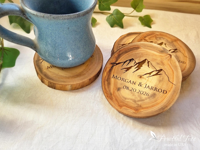 May include: Round wooden coasters with natural bark edges, featuring a laser-engraved mountain range graphic and the text MORGAN & JARROD 08.20.2026. A blue ceramic mug sits on one coaster, while two additional coasters are stacked nearby.