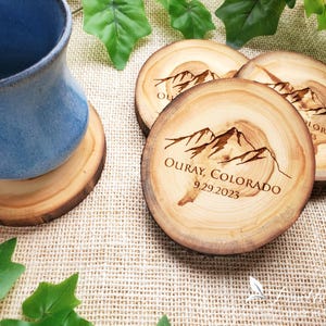 May include: Three round wooden coasters with natural bark edges, each featuring a laser-engraved mountain range graphic and the text "OURAY, COLORADO 9.29.2023". One coaster supports a blue ceramic mug, while the others are arranged nearby.
