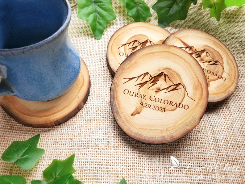 May include: Three round wooden coasters with natural bark edges, each featuring a laser-engraved mountain range graphic and the text "OURAY, COLORADO 9.29.2023". One coaster supports a blue ceramic mug, while the others are arranged nearby.