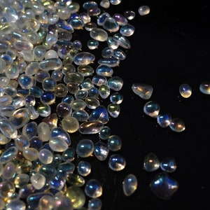 May include: A collection of iridescent glass beads in assorted sizes and forms. The beads are clear with a shimmering, multi-coloured coating that reflects light, producing a rainbow effect. The beads are scattered on a dark surface.