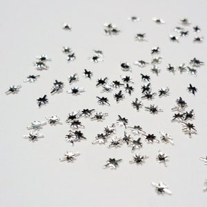 May include: Silver star-shaped embellishments scattered on a white background. These small, metallic stars are perfect for adding a touch of sparkle to crafts, nail art, or other projects.