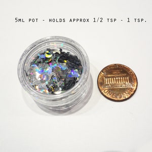 K&ouml;nnte beinhalten: Ein kleines, klares Plastikglas gef&uuml;llt mit schimmerndem silbernen und schwarzen Glitter. Das Glas ist etwa so gro&szlig; wie ein 1-Cent-St&uuml;ck.