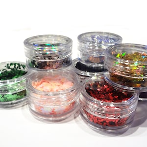 Pode incluir: Conjunto de pequenos frascos cheios de glitter colorido. O glitter é de várias formas e tamanhos, incluindo estrelas, corações e círculos. Os frascos são de plástico transparente e têm tampas prateadas.