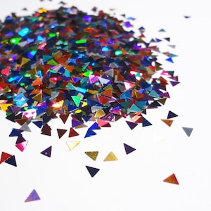May include: A collection of iridescent, triangular glitter pieces in various colours, including blue, green, red, and gold. The glitter is scattered on a white surface, creating a shimmering effect.