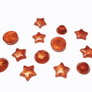 K&ouml;nnte beinhalten: Sortiment aus kupferfarbenen Harz-Cabochons in verschiedenen Formen, darunter Sterne und Kreise.