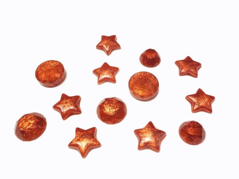K&ouml;nnte beinhalten: Sortiment aus kupferfarbenen Harz-Cabochons in verschiedenen Formen, darunter Sterne und Kreise.