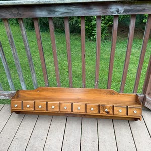 May include: A long, rectangular wooden wall shelf featuring ten small, aligned drawers. Each drawer is equipped with a single, small, round white knob. The shelf has a flat top surface and a slightly curved backboard, finished in a medium brown wood tone.