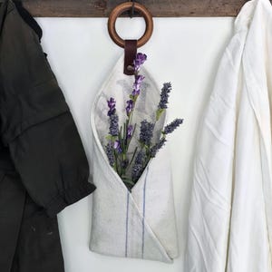 Pode incluir: Uma bolsa de linho branco com listras azuis, cheia de raminhos de lavanda roxos, pende de um anel de madeira e uma tira de couro. A bolsa está suspensa de uma viga de madeira rústica, com um casaco verde escuro e uma jaqueta branca de cada lado.