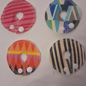 May include: Four colorful fabric nursing pads. One is pink with red stripes, another has a geometric pattern with blue and green, a third has an orange and pink design, and the last has black and beige stripes. Each pad has a snap closure.