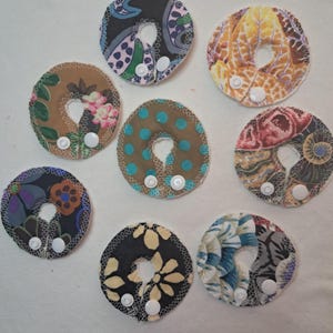 May include: A collection of eight fabric nursing pads with various patterns and colors. Each pad is circular with a central opening and features a snap closure. The pads display floral, polka dot, and abstract designs in shades of blue, brown, and yellow.
