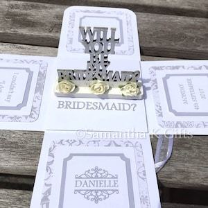 May include: A white box with a pop-up card that says "Will you be my bridesmaid?" with white flowers and a silver frame. The box also has a silver frame with the name "Danielle" inside.