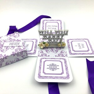 May include: A purple and white explosion box with a pop-up that says "Will You Marry Me?" The box is decorated with a damask pattern and has a purple ribbon.