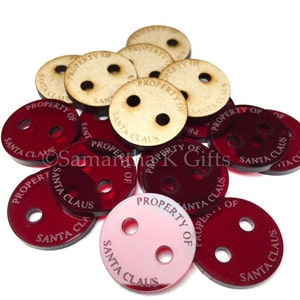 May include: A close-up of red and natural wood round ornaments with the text "Property of Santa Claus" engraved on them. The ornaments have three holes each.
