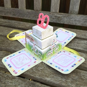 May include: A pink and white layered cake with a pink 80 on top. The cake is in a box with a yellow ribbon. The box has a green and white design with a sign that says "Tarka Trail". The box also has a pink and white design with the text "Life is measured by memories".