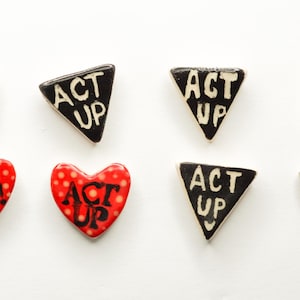 May include: An assortment of handmade pins and brooches. The collection includes black triangular pins with "ACT UP" in white lettering, and red heart-shaped pins with the same text. The hearts have white polka dots.