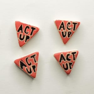 May include: Six triangular, red-orange ceramic tiles with the words "ACT UP" in black lettering. The tiles are arranged on a white surface. The tiles are likely decorative or part of a larger design.