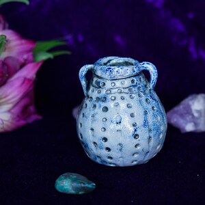 May include: A small, blue and white ceramic jar with a textured surface and two handles. The jar is sitting on a purple velvet surface.