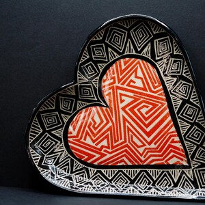 May include: A heart-shaped ceramic dish with a black and white geometric pattern around the edges and a red and white geometric pattern in the center.