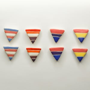 May include: Eight colorful, triangular ceramic pieces arranged on a white surface. Each piece features a unique striped pattern in shades of red, blue, orange, purple, and yellow. The geometric shapes are evenly spaced.