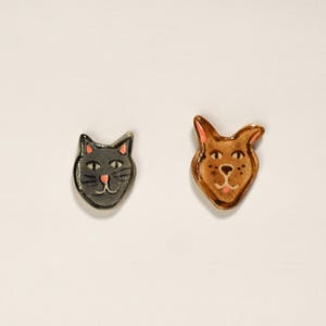 May include: Two ceramic cat head figurines. One is gray with pink accents, the other is brown with pink accents. Both have black whiskers and are set against a white background.