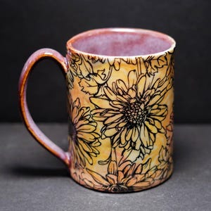May include: A ceramic mug with a floral design. The mug features a yellow and orange background with black floral outlines. The interior and handle have a purple and brown gradient. The mug is designed for hot beverages.