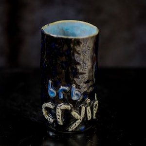 May include: A black ceramic mug with a blue and white glaze. The mug has the words "brb" and "cry" written on it in white.