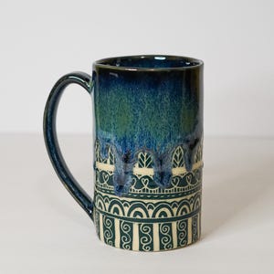 May include: A tall ceramic mug featuring a glossy dark blue and green drip glaze on the upper portion. The lower section is cream-colored with dark blue hand-drawn patterns including horizontal lines, heart shapes, arches, and vertical swirls.