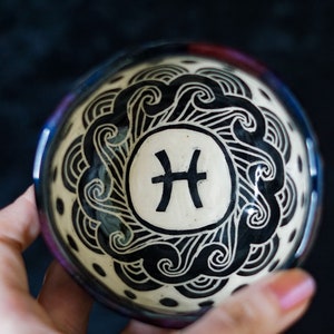 May include: A small white ceramic bowl with a black and white design of waves and the Pisces zodiac symbol. The bowl has a black rim and a speckled glaze.