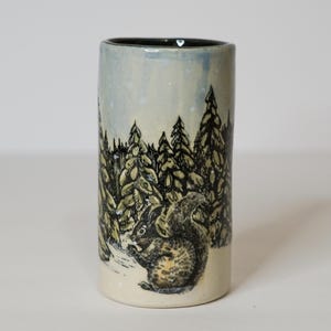 May include: A cylindrical ceramic vase featuring a hand-drawn black ink illustration of a squirrel sitting in a forest of pine trees. The background has a light blue and white wash, while the interior of the vessel is a dark charcoal gray.