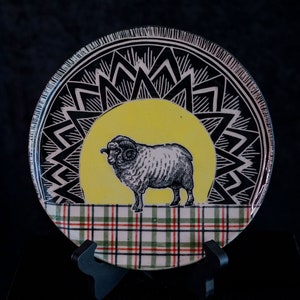 May include: A decorative ceramic plate featuring a detailed illustration of a ram against a yellow backdrop. The plate has a black and white geometric border and a plaid pattern at the bottom. The plate is displayed on a black stand.
