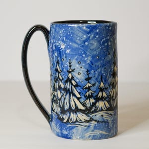 May include: A tall ceramic mug with a glossy black handle and a cylindrical body. The exterior features a hand-painted winter scene with white pine trees against a textured blue background, accented with white dots representing falling snow.