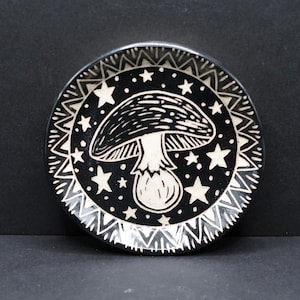May include: A black and white decorative plate featuring a mushroom design. The plate has a white star and geometric pattern around the edge. The mushroom is in the center of the plate.