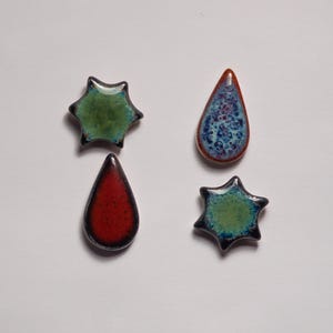 May include: Four ceramic decorative pieces in star and teardrop shapes. The star shapes are green with dark blue edges. One teardrop is red with a black edge, and the other is blue with a brown edge. These pieces are likely for jewelry or craft projects.