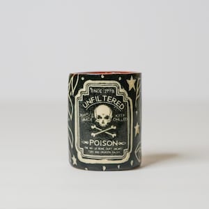 May include: A black ceramic mug with an orange interior featuring a white graphic label that reads UNFILTERED POISON SINCE 1778, with a skull and crossbones illustration, stars, and additional text detailing ingredients like bone dust and dragon scale.