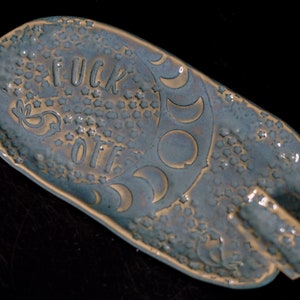 May include: A blue ceramic dish with a textured surface and a gold design. The dish has a small handle on the side and the words "F**K OFF" are written in gold on the surface.