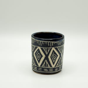 May include: A cylindrical ceramic cup with a dark blue exterior and a cream-colored geometric pattern. The pattern consists of diamond shapes and lines. The interior of the cup is dark blue.