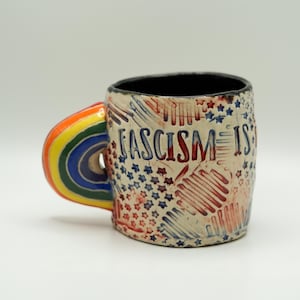 May include: A ceramic mug with a rainbow handle and the words "FASCISM IS" in red and blue. The mug's body is off-white with red and blue star and stripe designs. The interior of the mug is black.
